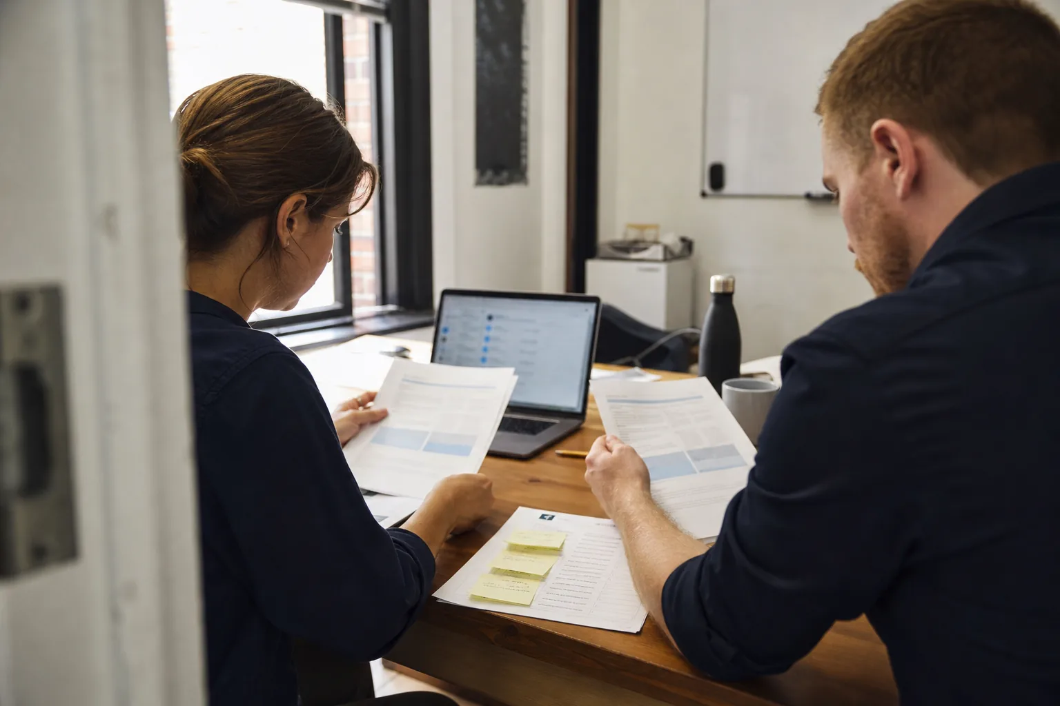 Two staff at a gov-tech company comparing printed compliance software proposals next to an agency security questionnaire
